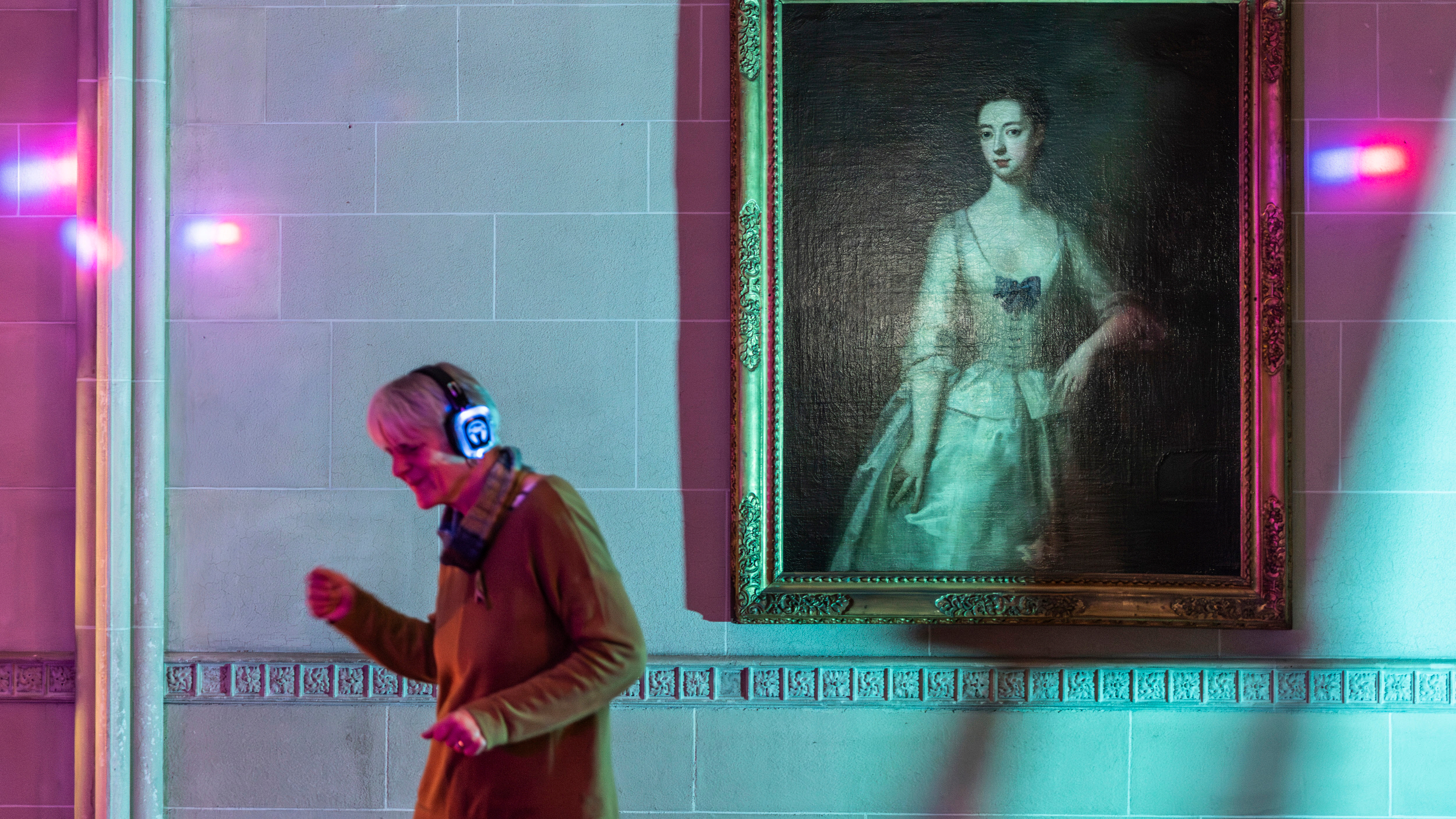 A person wearing headphones dances beside a large portrait painting inside a gallery lit with colourful lights.