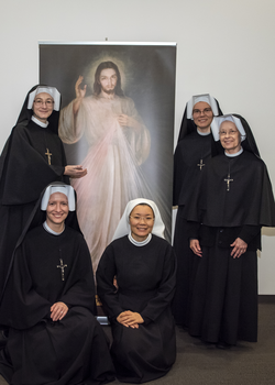 5 Catholic nuns dressed in habits adorned with crosses kneel and stand next to a poster of Jesus Christ against a bare wall.