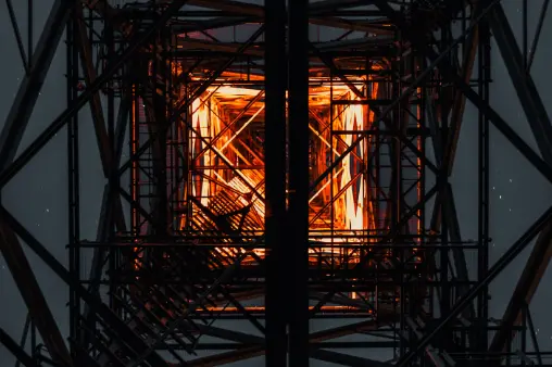 An abstract, low-angle shot looking up through the metal framework of a large tower or pylon at night, with a bright orange light illuminating the structure from within.