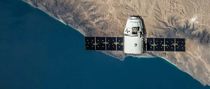 A white satellite with long, dark solar panels orbits above a coastline, with deep blue ocean meeting tan, arid land. The satellite has a "SPACEX" logo visible.