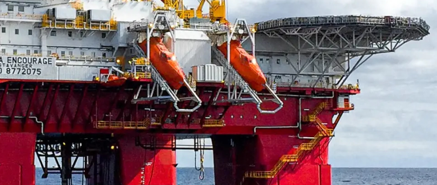 Close-up of a large red and white offshore oil rig or drilling platform, showing two orange enclosed lifeboats suspended from the upper deck.