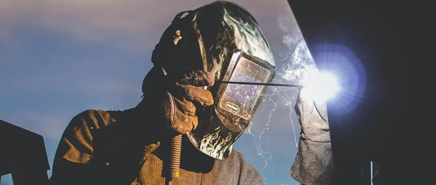 A worker wearing a welding mask and heavy gloves performs arc welding outdoors, with bright sparks and light emanating from the weld against a bright sky.