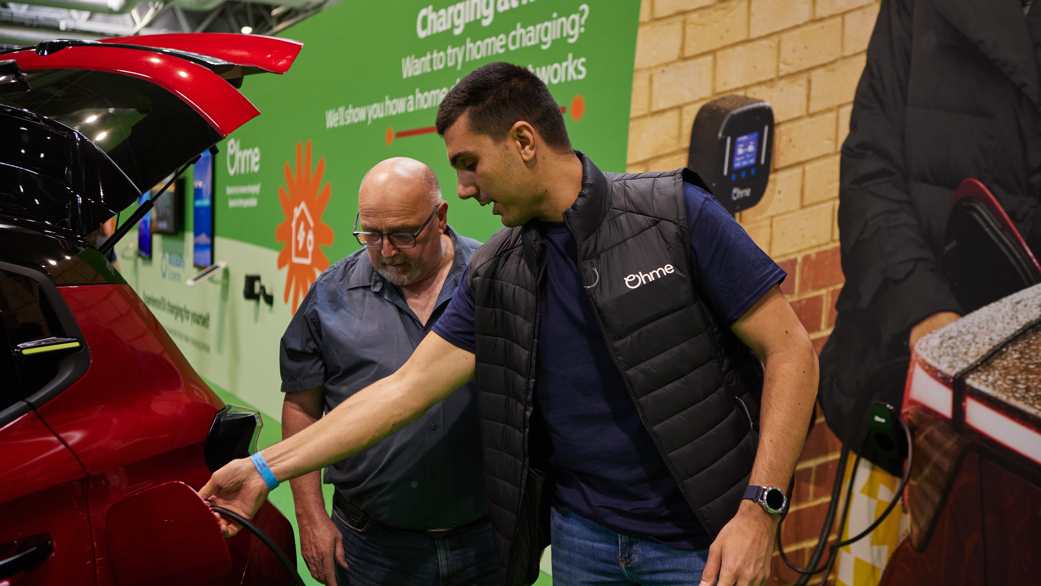 An Ohme specialist plugs a charging cable into a red electric car while another man watches at Motability Scheme Live in Birmingham. They are at a stand about home charging.