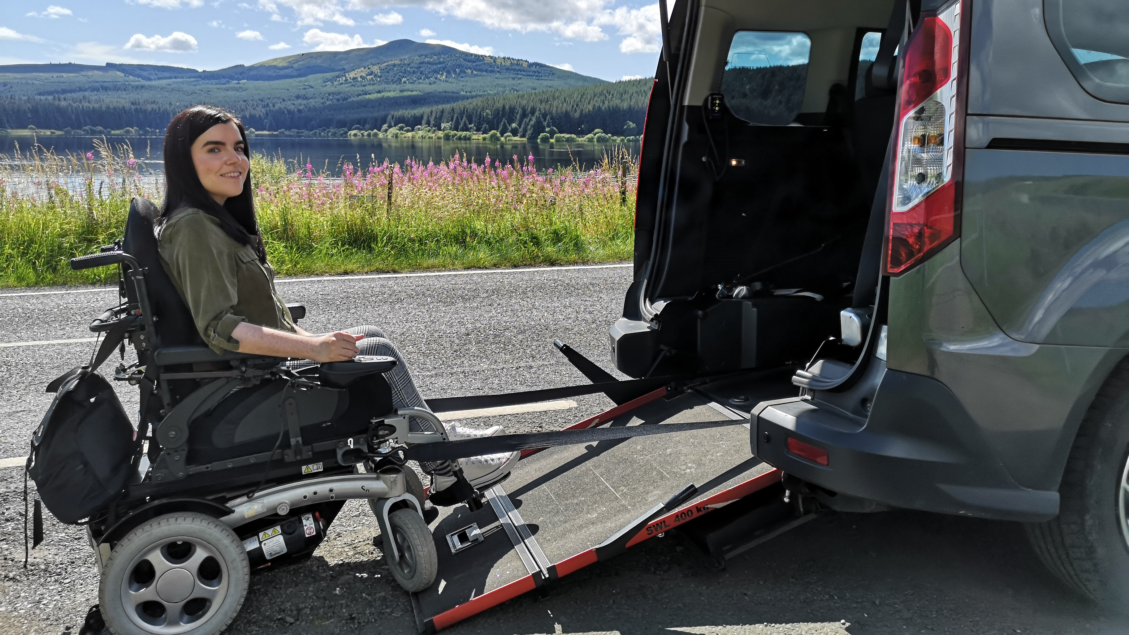 Emma Muldoon sits in a powered wheelchair beside a Wheelchair Accessible Vehicle (WAV) with a rear ramp deployed, parked by a lake with hills and trees in the background on a sunny day.