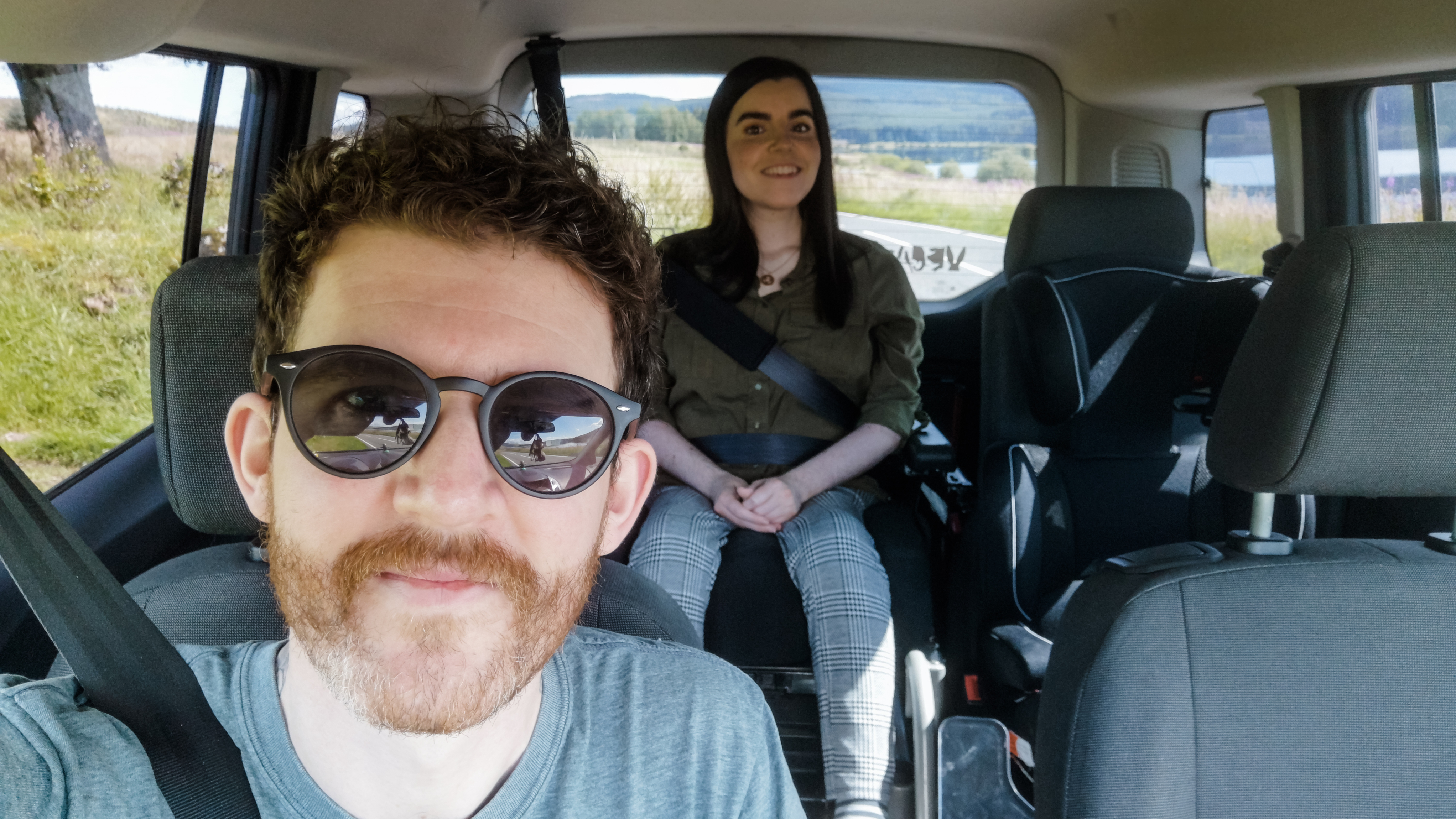 Emma Muldoon sits in her powered wheelchair at the base of a rear access ramp on a parked Wheelchair Accessible Vehicle (WAV) while a young child stands on the ramp beside her, with trees and a fence in the background.