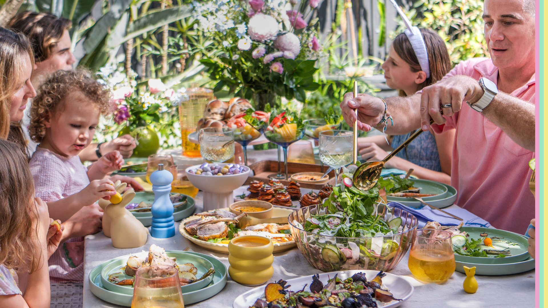 A family gathered around an outdoor table sharing an Easter meal, with plates of salads, roast vegetables and seafood, drinks in pastel glasses, and festive table décor set among greenery.