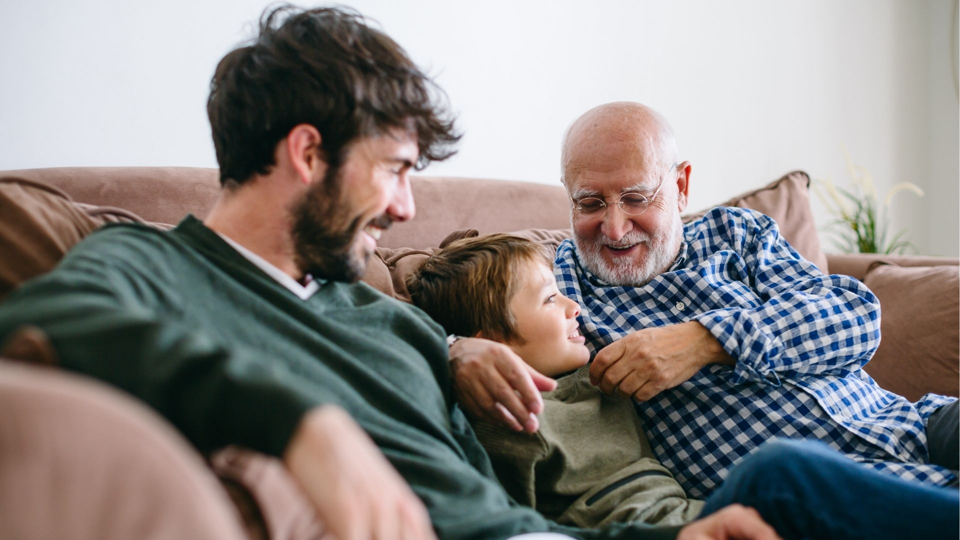 Granddad and Dad relaxing on a sofa with a child, spending quality time together. Dad sharing a hug with two children while holding a Father’s Day gift. ad receiving a wrapped Father’s Day gift from a child while sharing a meal together