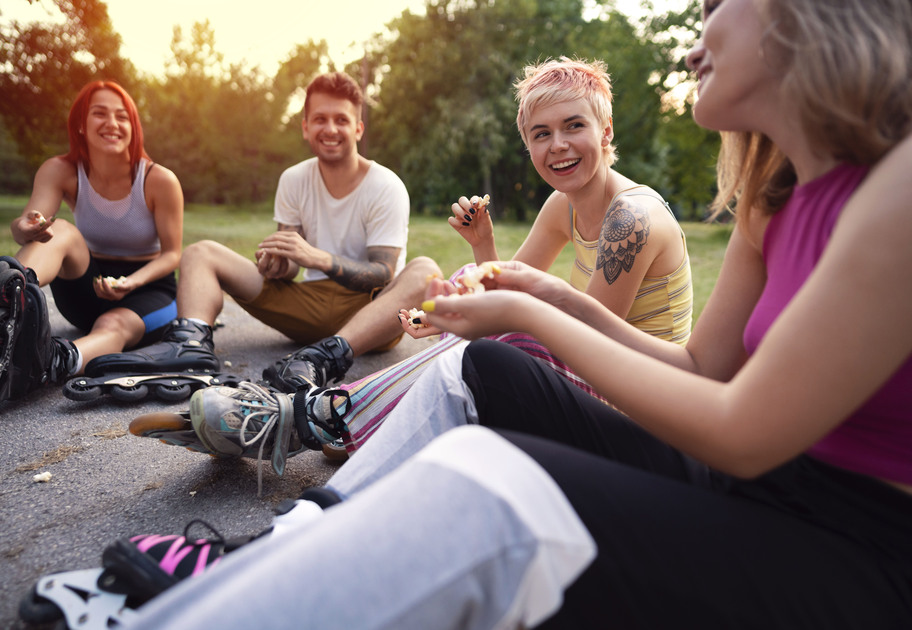 Four friends in rollerblades sitting on a park path, laughing and sharing snacks at sunset.