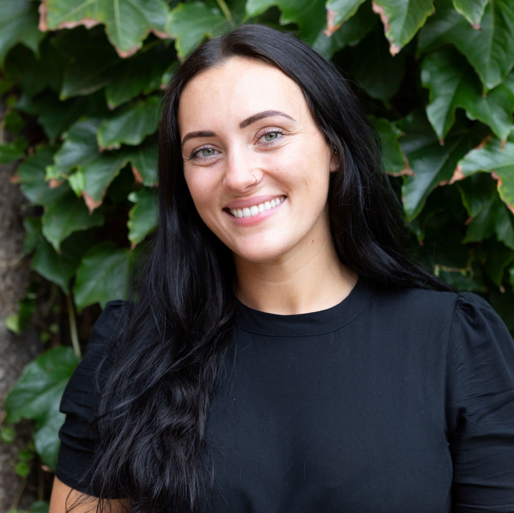 A portrait of a smiling woman with long, dark hair and light-colored eyes. She is wearing a black top and standing in front of a wall covered in lush green ivy.