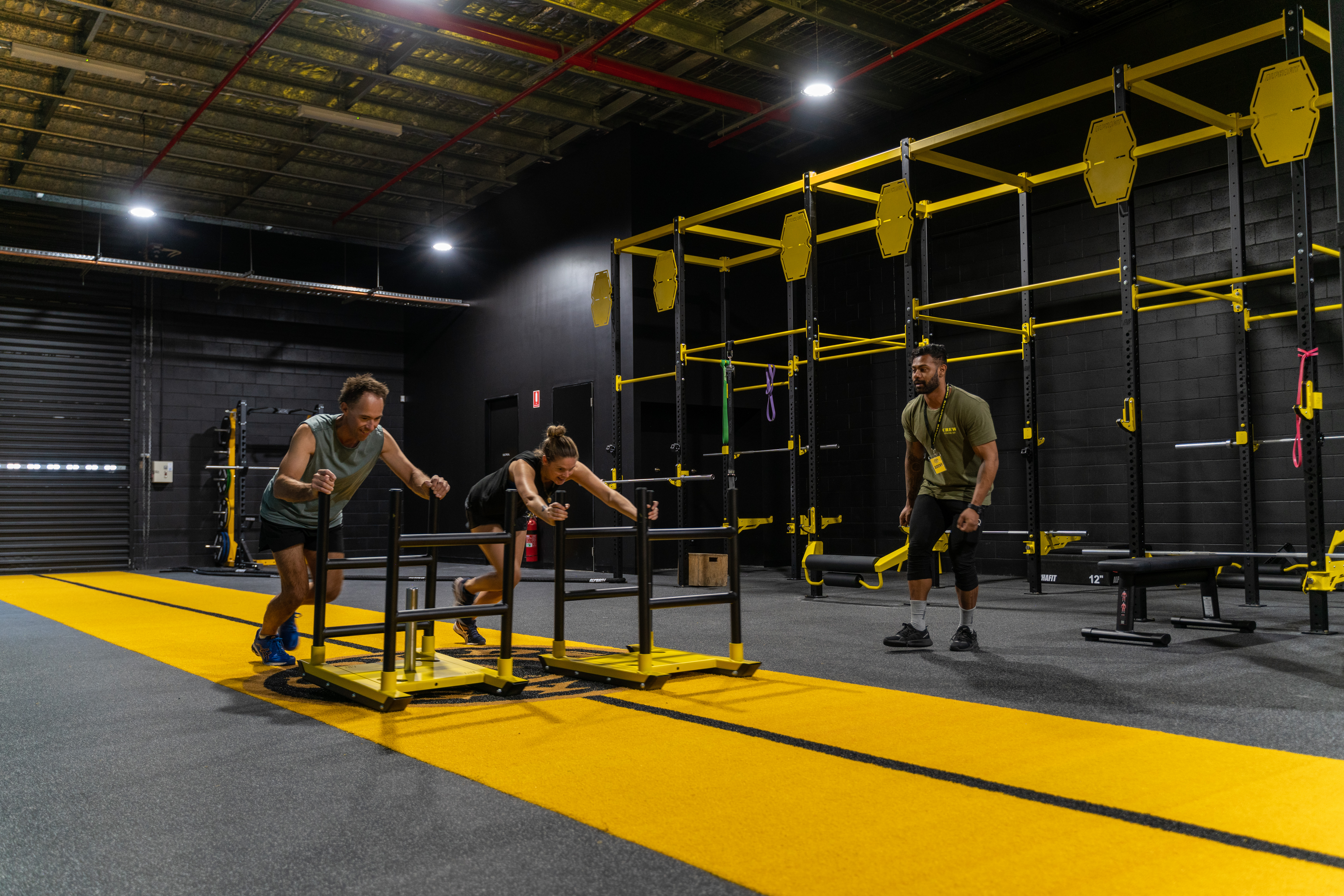 2 people in a gym pushing weights along the ground with their trainer encouraging them along.