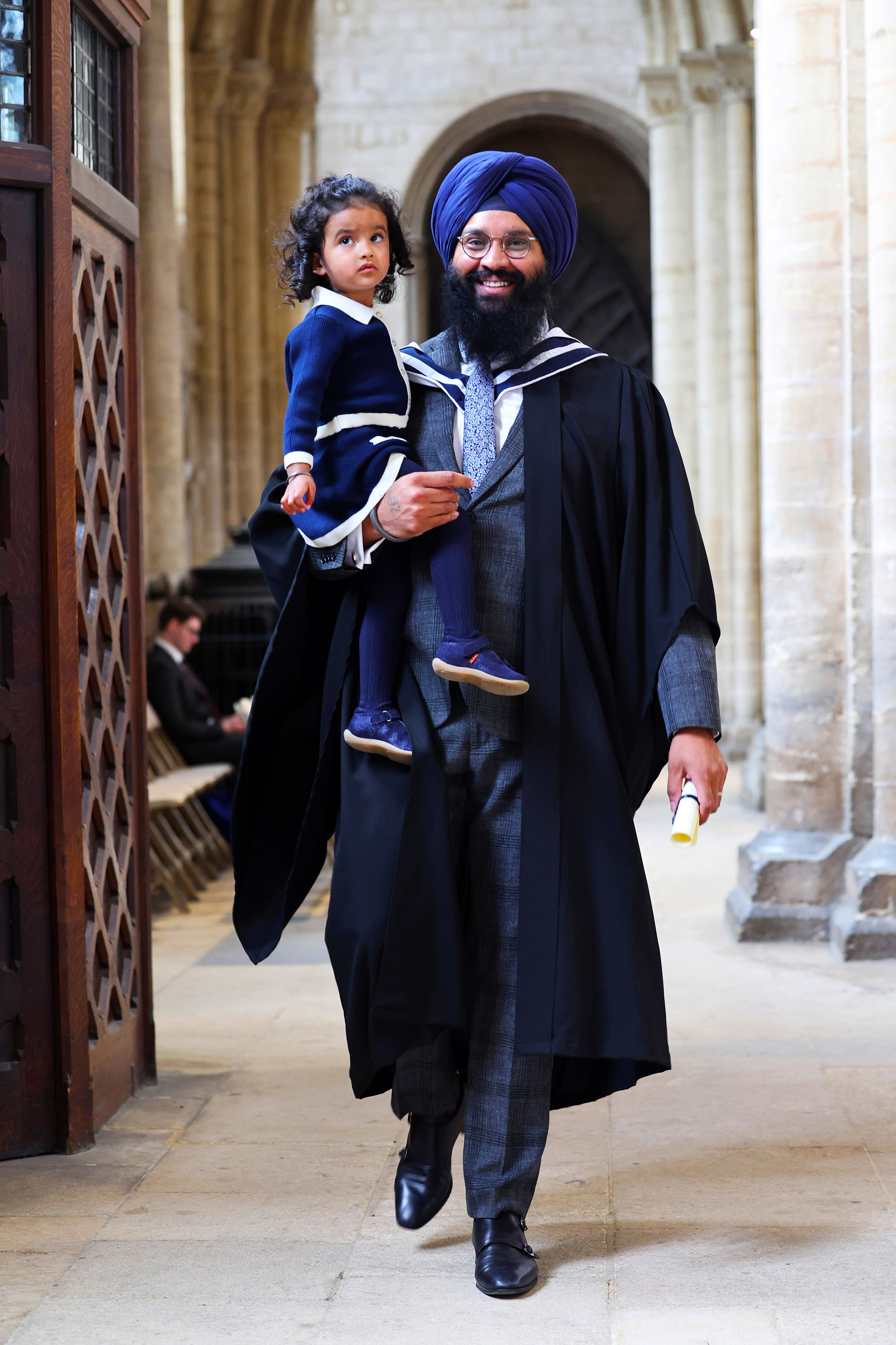 Graduate holding a young child and wearing a turban inside a hall