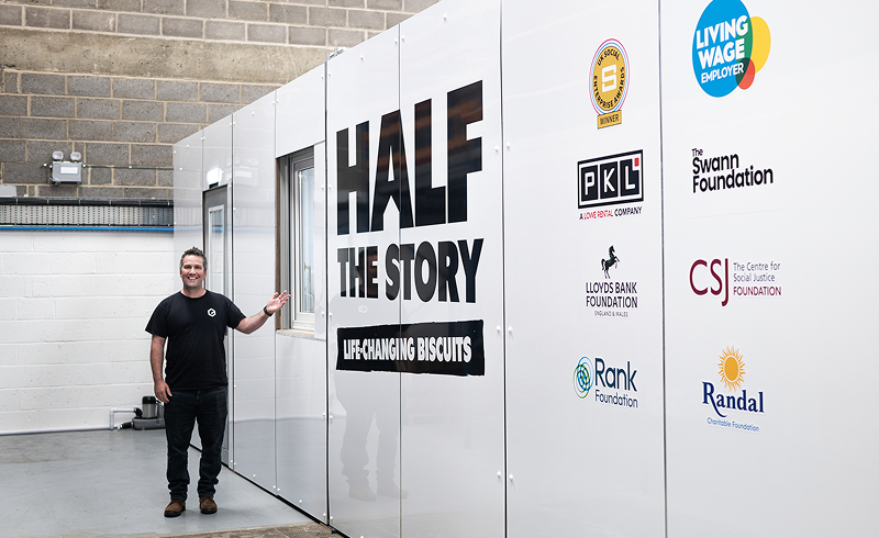 Smiling man standing next to a wall with a large 'half the story' logo on it, he's pointing to the wall