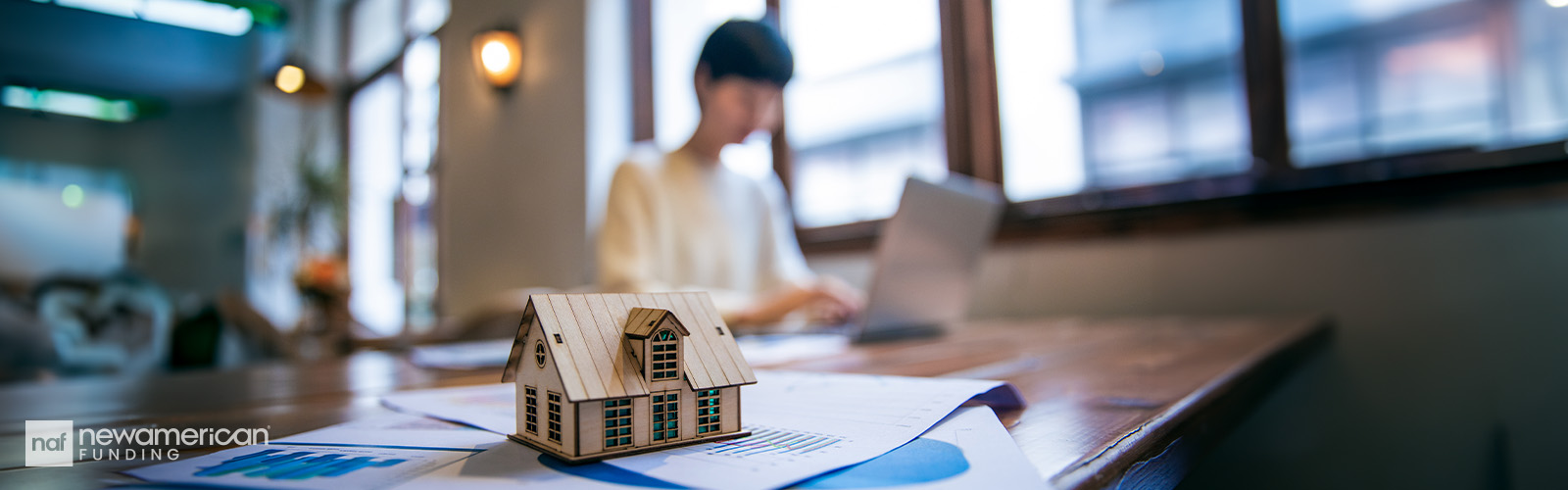 Someone is working on a laptop next to a stack of paperwork with a home on top of it.