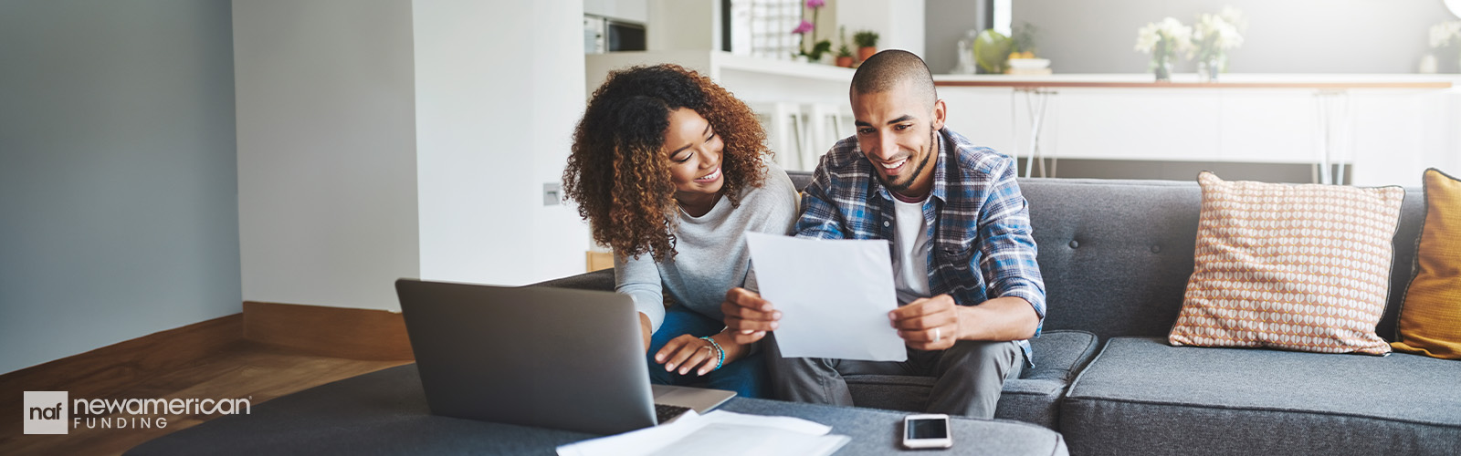 A Black man and Black woman sit together on a couch in a nice living room. They are smiling at a piece of paper the man is holding.  
