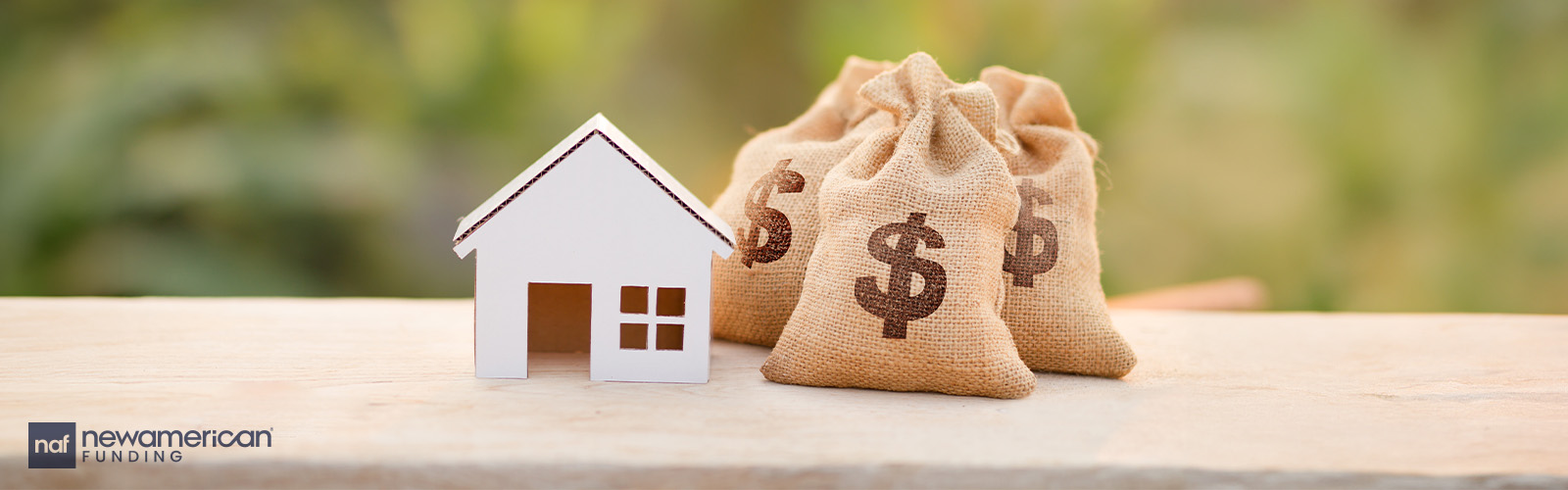 A small wooden model of a house and two little bags with a money symbol on them sit on a desk top 