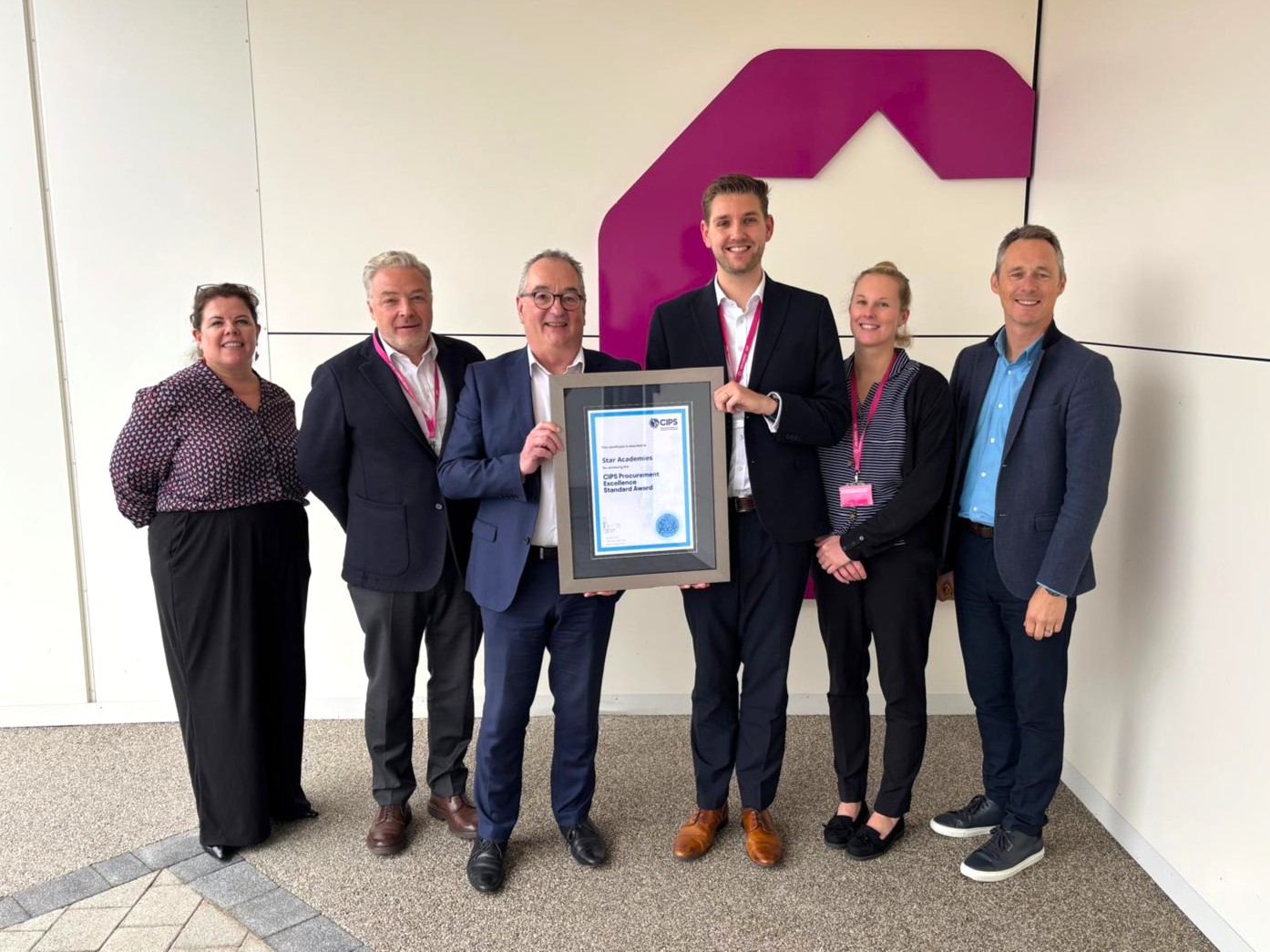 Group of 4 men and 2 women gathered. Two men in the centre hold up a frame that contains the CIPS Procurement Excellence Standard Award, The background shows the logo of Star Academies