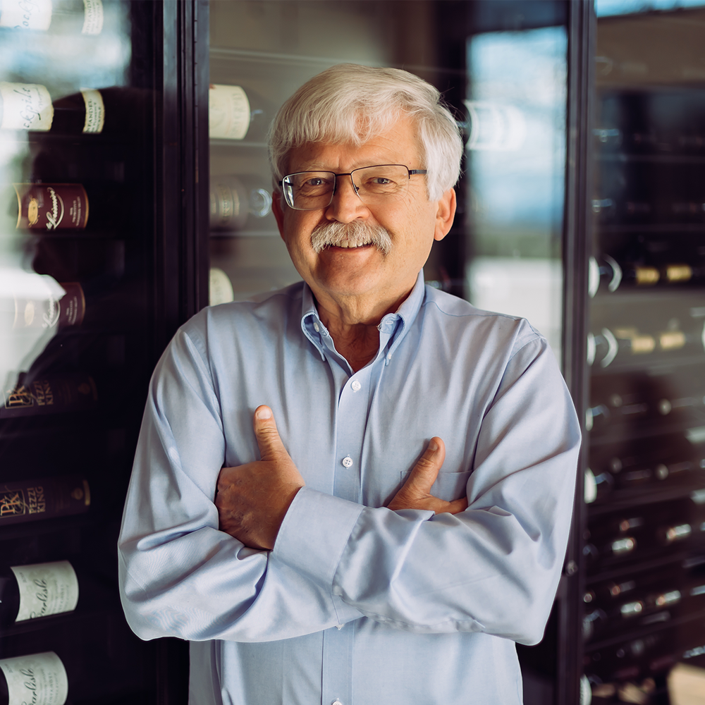 A photograph of a smiling man with white hair, a mustache, and glasses. He is wearing a light blue button-down shirt and has his arms crossed while leaning against a large, modern glass wine cellar filled with bottles.