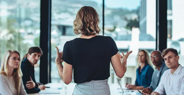 Back of a standing woman gesturing with her hands to people sat at a large meeting table in an office setting