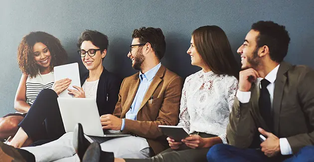 Five people sat in a row next to each other, all wearing smart clothing and looking at the 2nd person who is holding papers