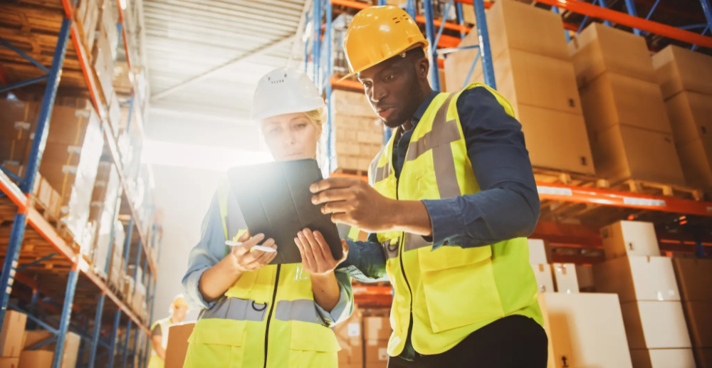 A man and a woman wearing hi vis jackets and helmets, looking together at an iPad in a warehouse