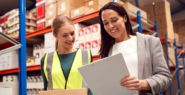 Two women in a warehouse looking at a piece of paper that one of them is holding, the other is wearing a hi vis jacket and is holding a box