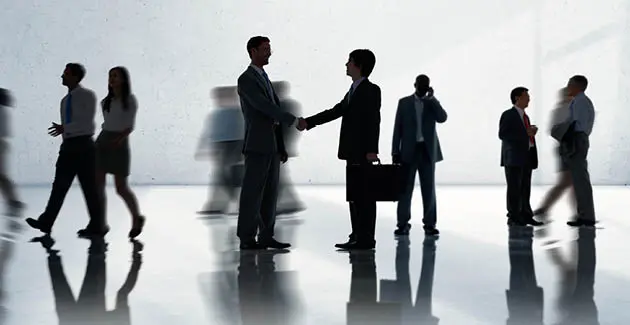 A silhouetted style photo of two men wearing business suits shaking hands, more professionals are in the background, some are blurred 