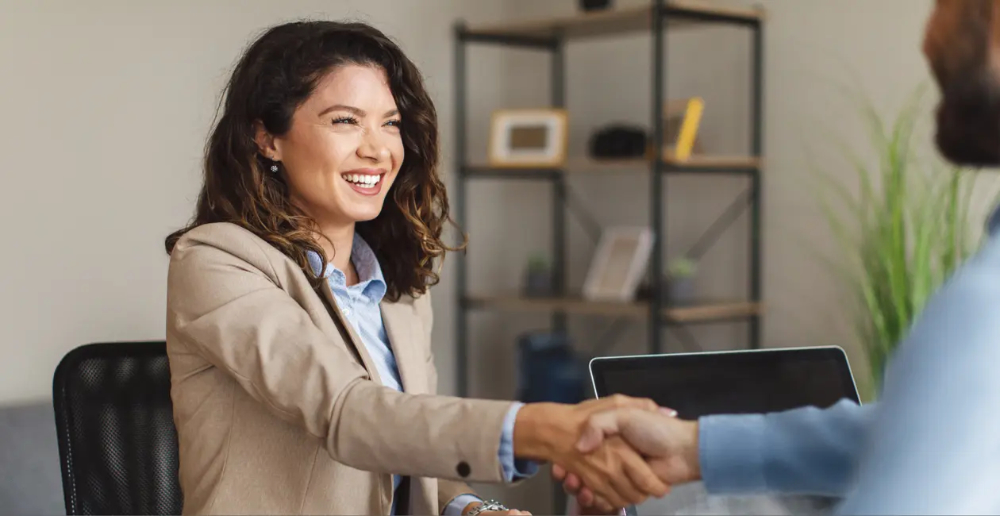 A woman shaking hands with a man that is mostly out of frame, she is wearing a beige suit jacket and is smiling