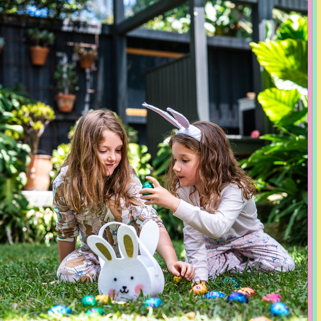 Two children in Easter themed pyjamas, one wearing bunny ears, are sitting on grass examining colorful Easter eggs near a bunny-shaped basket. Lush plants and hanging pots are in the background.
