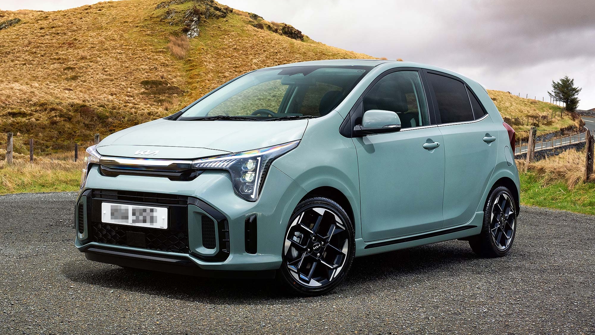 Light green Kia Picanto parked on a roadside with grassy hills and cloudy sky, available on the Motability Scheme with no Advance Payment.