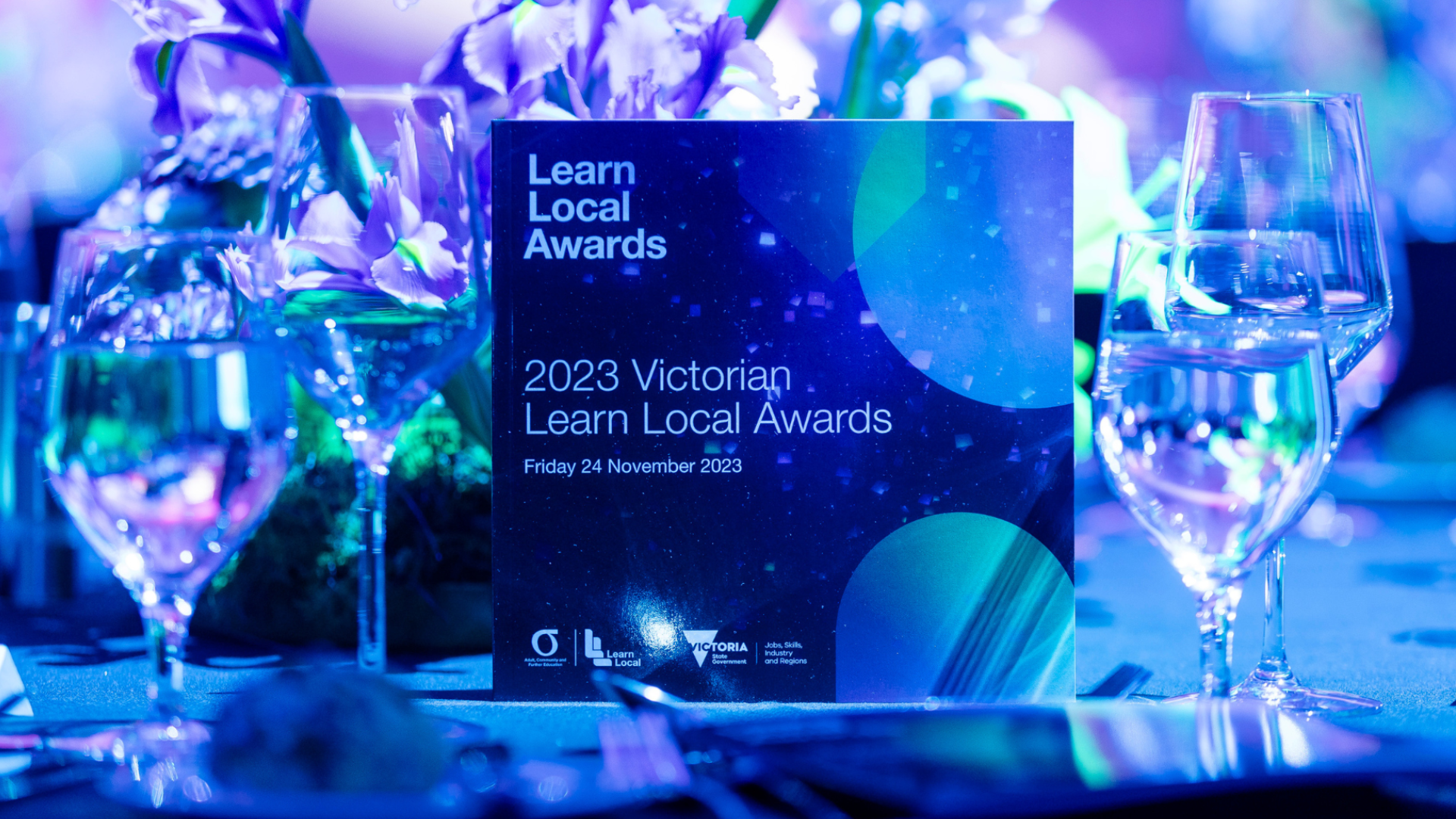 Award drowned in blue and purple lighting, placed on a dining table surrounded by wine glasses filled with a clear substance, in the background sits a floral centerpiece consisting of flowers with purple petals.