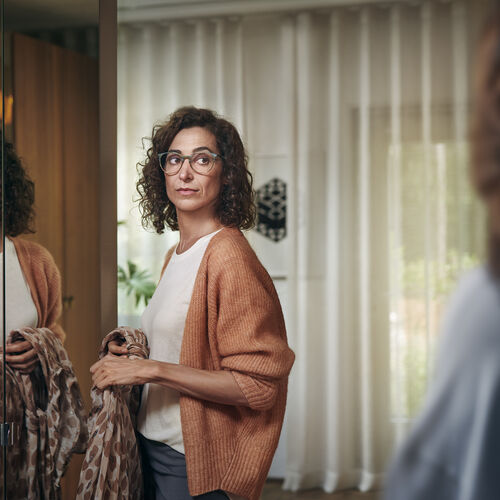 穿著橘色羊毛衫、戴眼鏡、在鏡子前拿著圍巾的女人