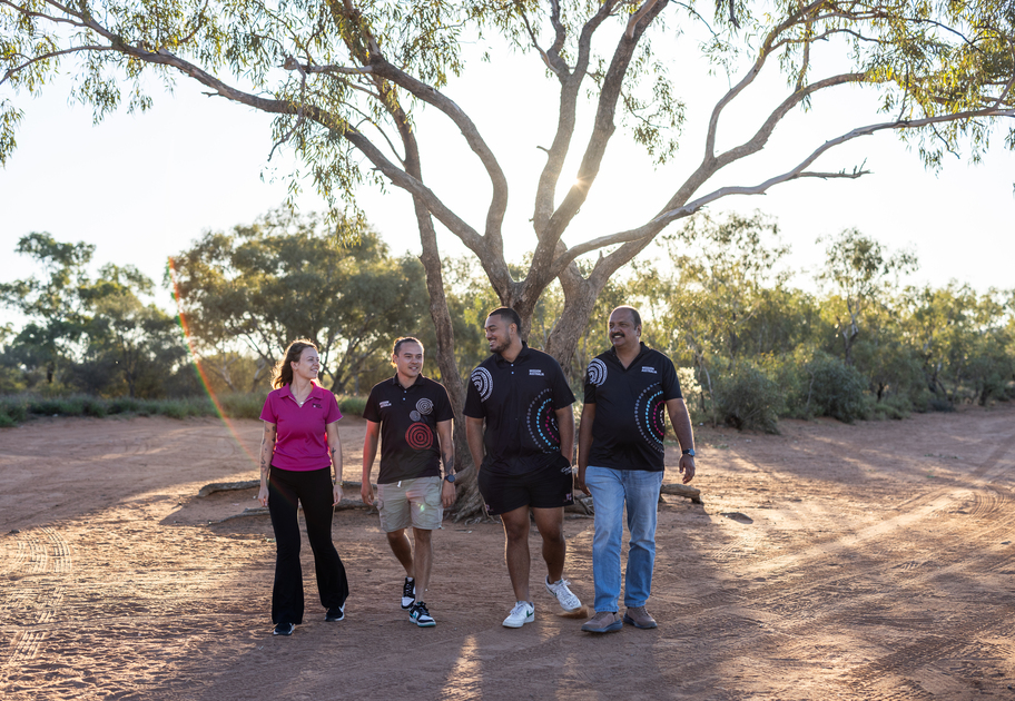 Four Mission Australia staff walking together on a red dirt track under a large tree in a rural outback setting.