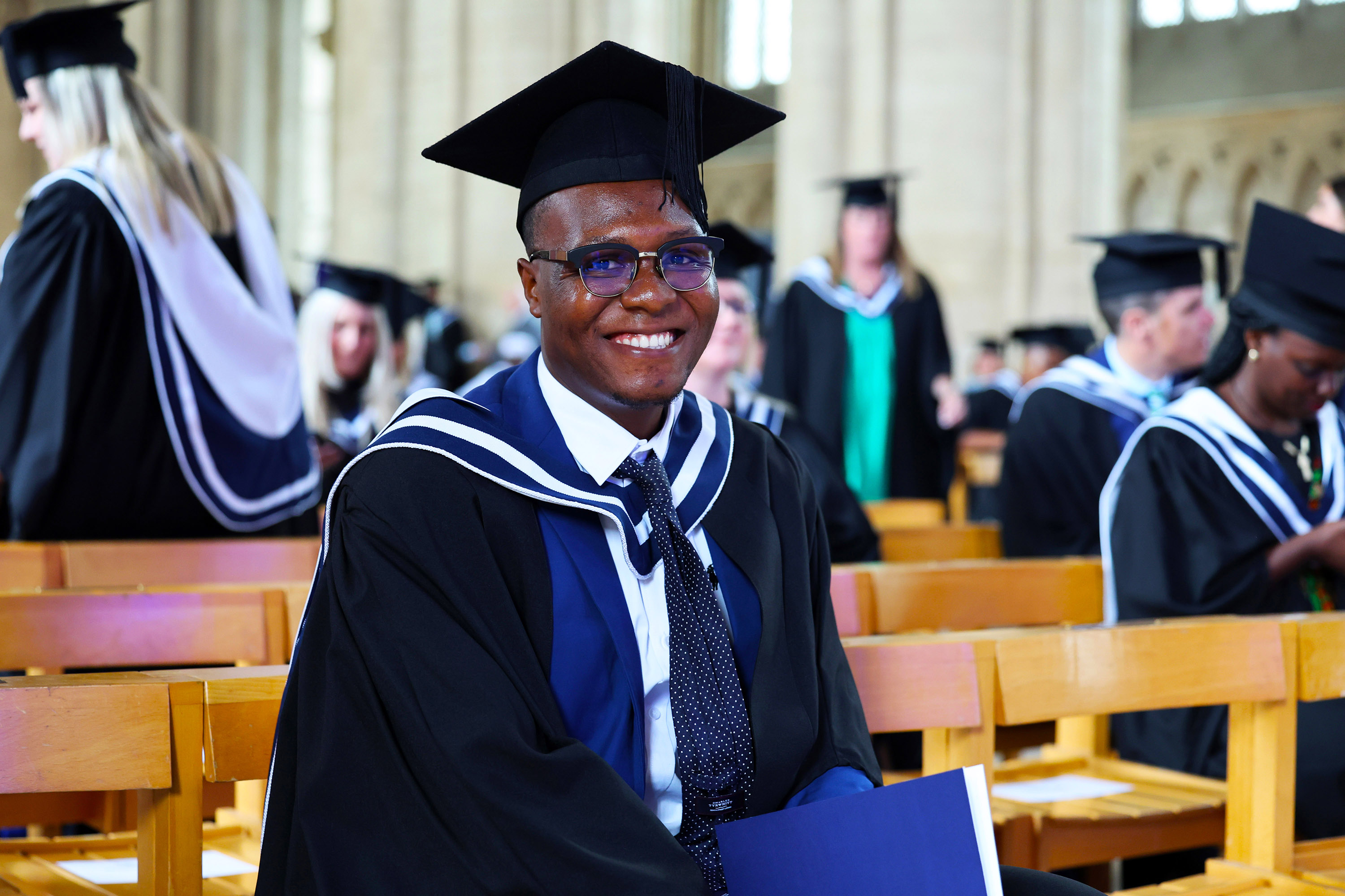 Male graduate with glasses smiling while seated on a wooden bench