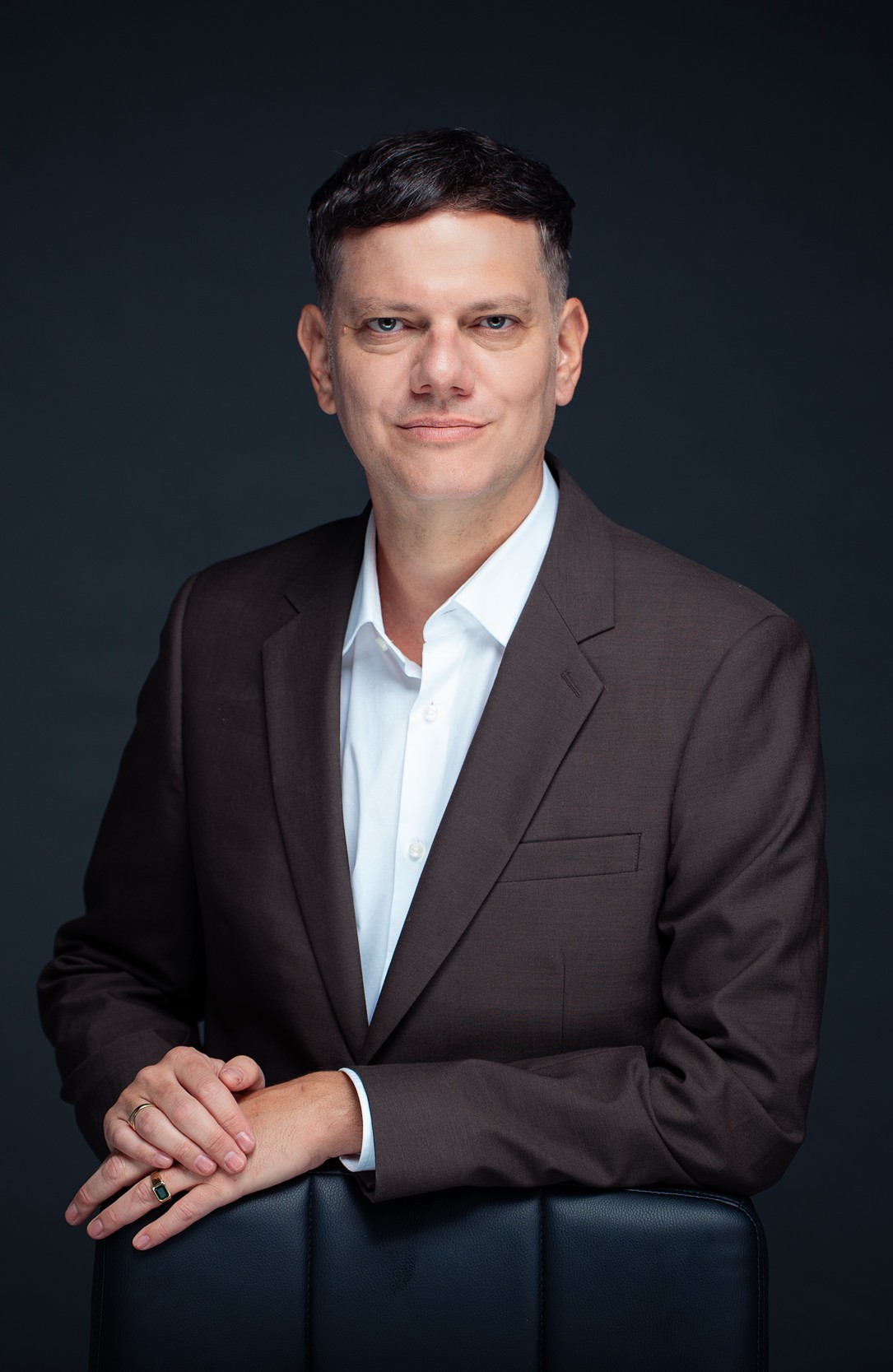 A medium-length, professional headshot of a man with dark, short hair and light eyes. He is wearing a dark brown blazer over a crisp white button-down shirt. He is standing behind a black leather chair, resting his crossed hands on the top of the chair's back. He has two rings on his fingersone a simple gold band and the other featuring a green rectangular stone. The background is a solid, dark slate gray.
