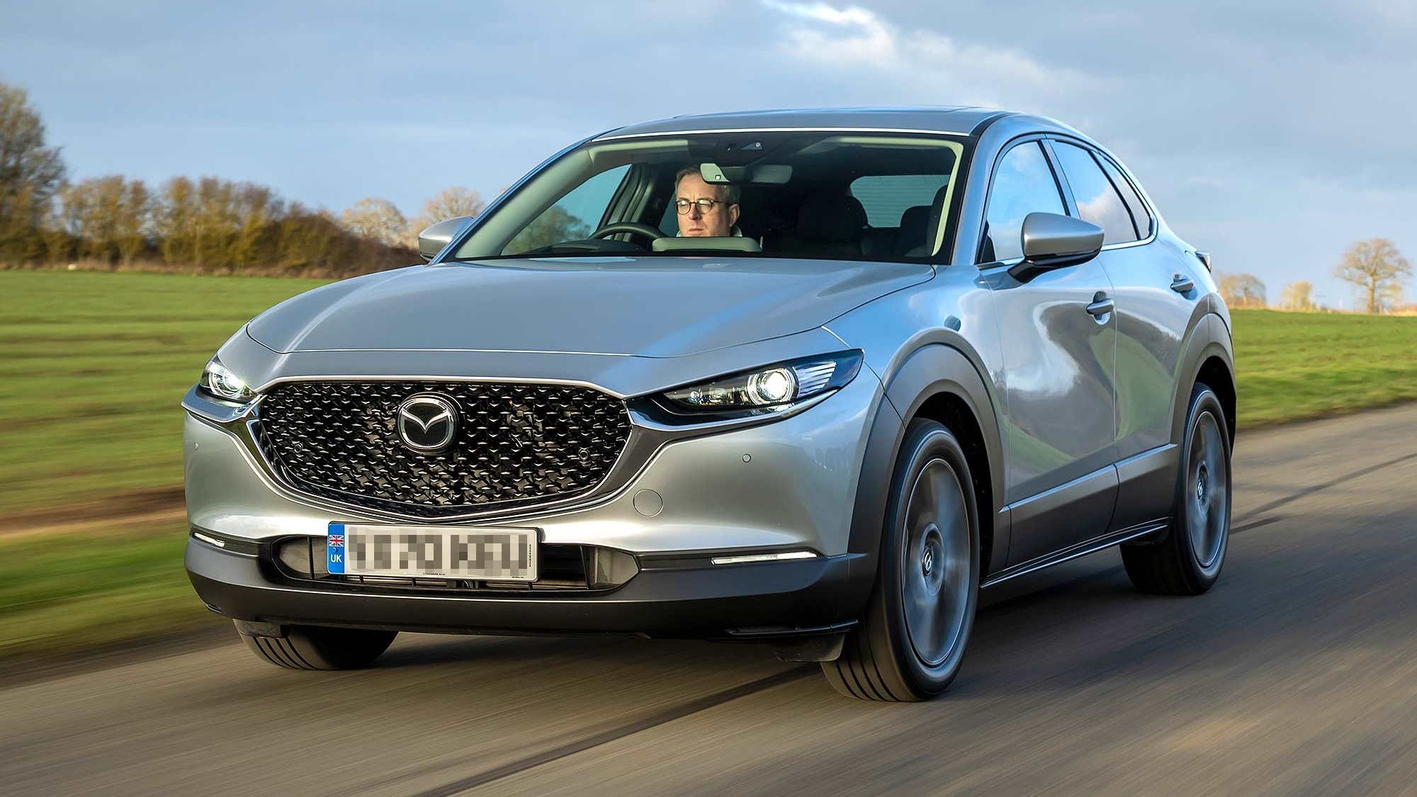 Silver Mazda CX 30 driving on a rural road, front three quarter view, with a driver visible through the windscreen.