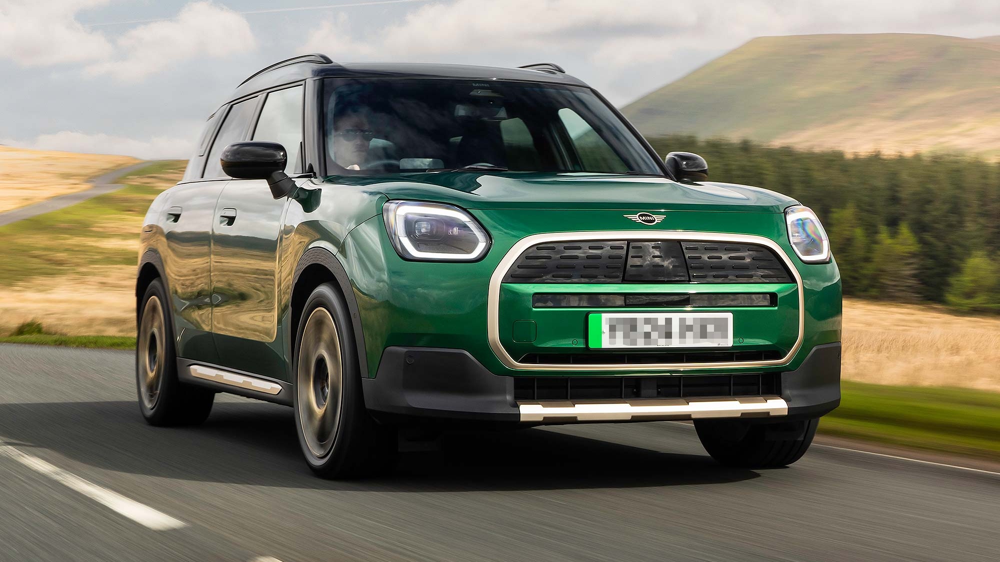 Green MINI Countryman SUV driving on an open countryside road under blue sky, available on the Motability Scheme with no Advance Payment.
