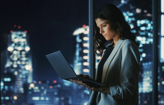 Woman standing in front of office windows at night working on laptop