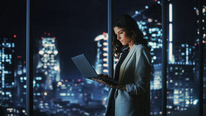 Woman standing in front of office windows at night working on laptop