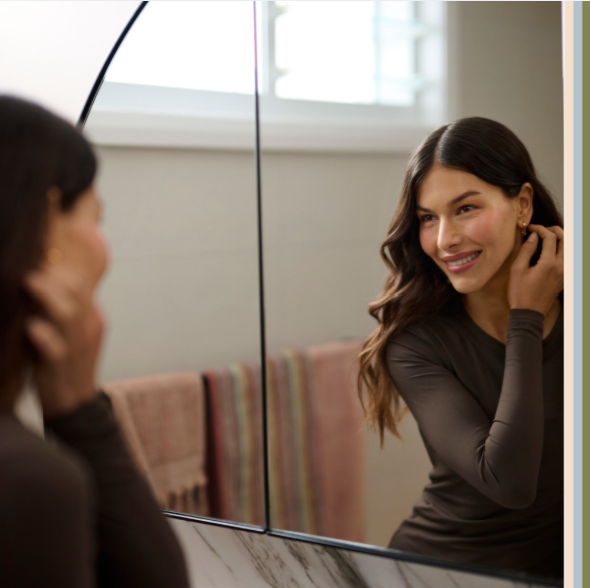 Woman getting ready in front of mirror wearing fitted top and adjusting earrings during beauty routine