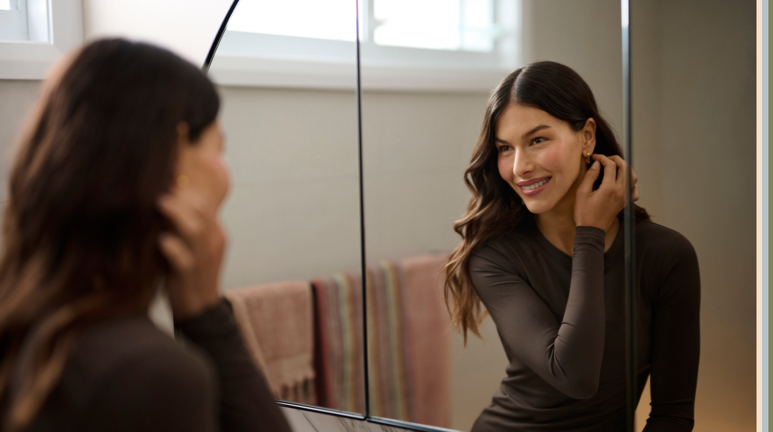 Woman getting ready in front of mirror wearing fitted top and adjusting earrings during beauty routine