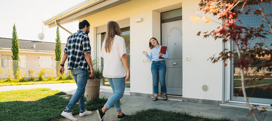 A woman with a clipboard welcoming a couple into the front door of a home.