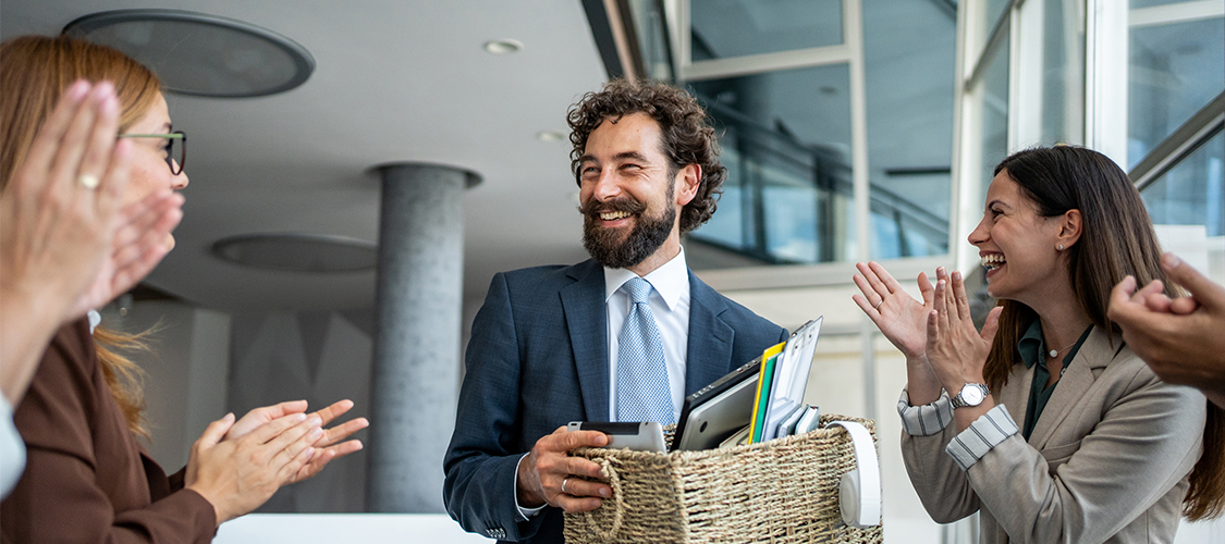 A man leaving an office with a box of belongings, smiling, surrounded by people clapping.