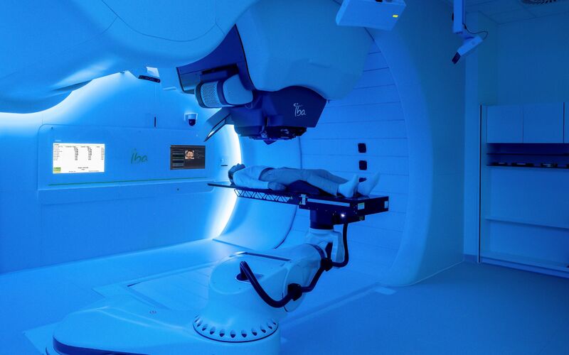 A photo image shows a proton therapy unit with a patient. The image is edited with a blue shade filter.