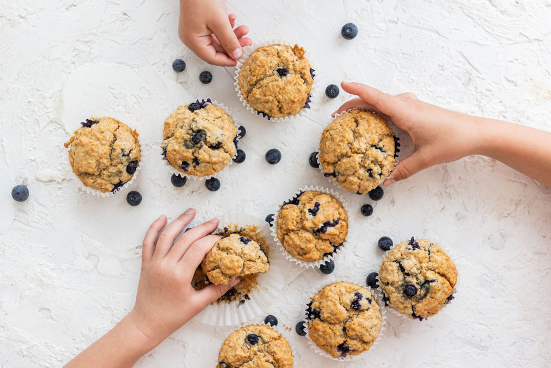 Nine homemade blueberry muffins are spread across a white kitchen benchtop, amongst scattered blueberries. Two sets of children’s hands are reaching for the muffins.