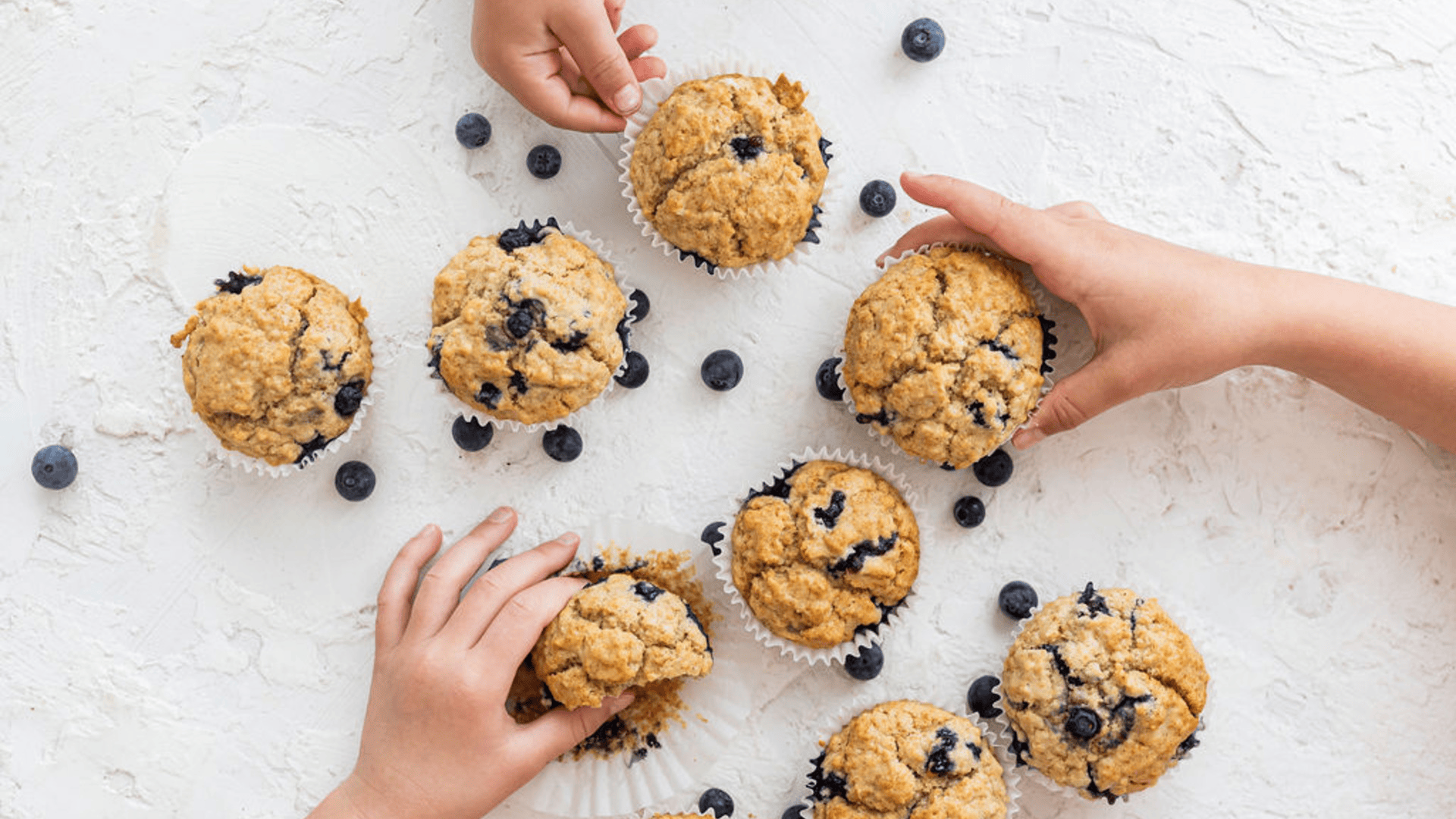 Nine homemade blueberry muffins are spread across a white kitchen benchtop, amongst scattered blueberries. Two sets of children’s hands are reaching for the muffins.