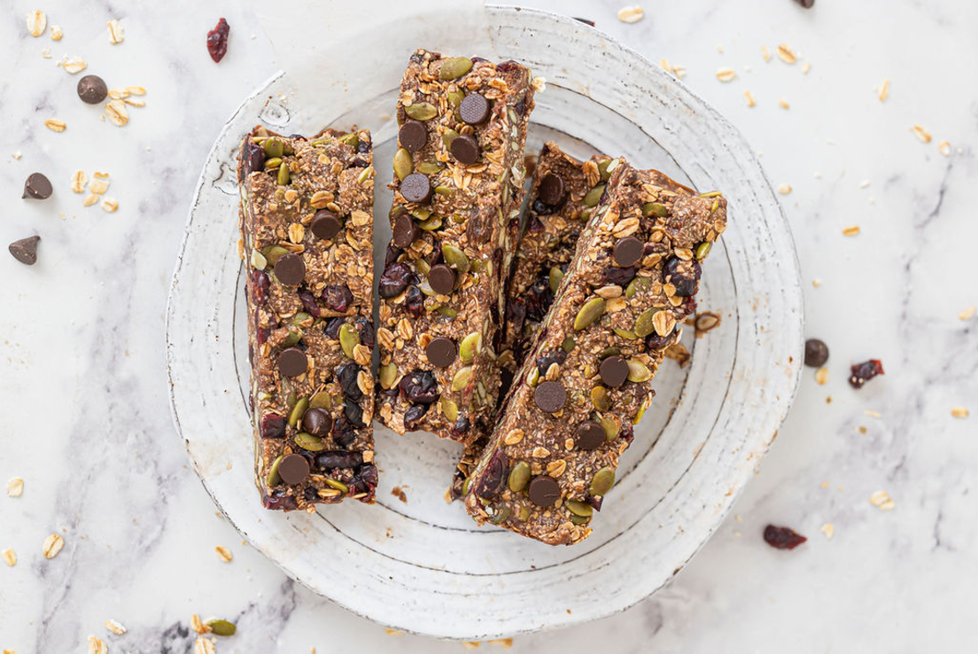 Three homemade granola bars are placed upon baking paper. The bars are sprinkled with pepitas, dark chocolate drops and shredded coconut.