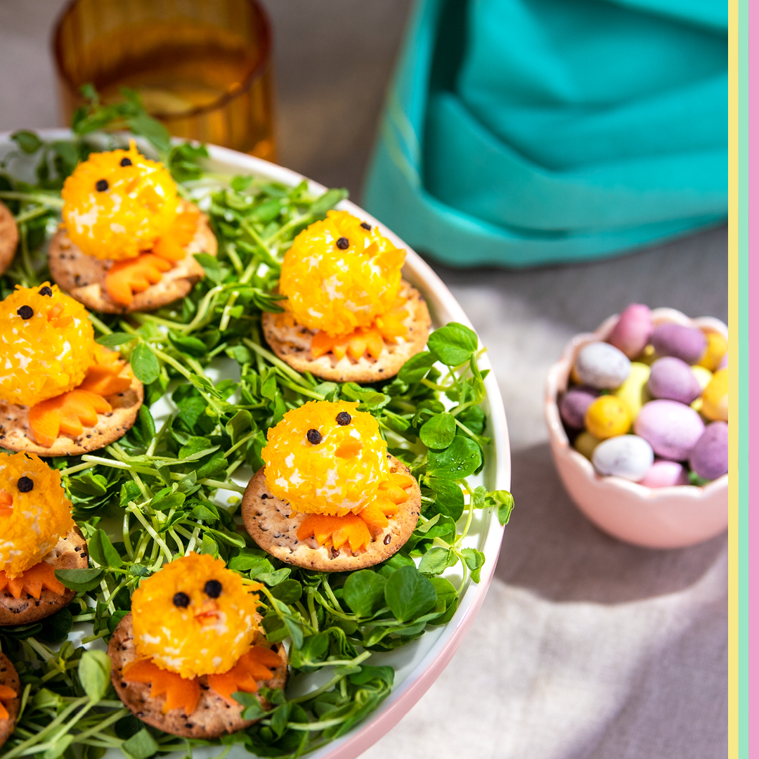 Mini cheese balls rolled in shredded cheese and decorated to look like baby chicks itting on a bed of herbs with sugar Easter eggs sitting behind 