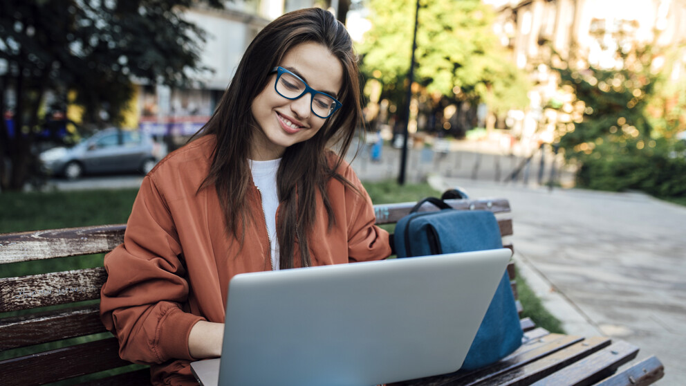 Mulher usando óculos azuis sorrindo para um laptop em um banco no parque