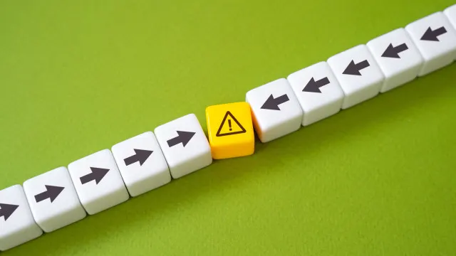 A row of white, cube-shaped blocks lies on a bright green surface. Most of the white blocks have a black arrow pointing to the left, indicating a sequence or flow. However, the four blocks on the left end have arrows pointing to the right.In the middle of the row, separating the right-pointing arrows from the left-pointing arrows, is a single yellow cube with a black exclamation mark inside a triangle (a warning symbol). The white blocks immediately next to the yellow block appear to be displaced slightly, as if the yellow block is a disruption or an obstacle that has caused the flow to change direction.