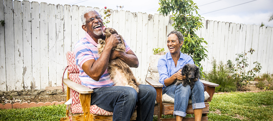 A senior Black woman and man sit laughing together and playing with dogs in their back yard